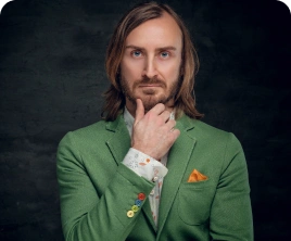 Vállig érő hajjal és szakállal, zöld öltönykabátban álló férfi gondolkodva pózol, állát kezére támasztva. Színes gombok és minták teszik különlegessé a ruhát